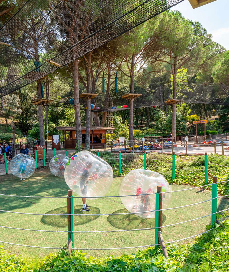 Niños y adultos jugando en burbujas inflables en un parque de actividades con pista de karts y circuito de aventura en los árboles al fondo.